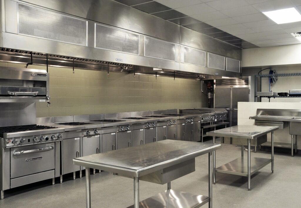 Industrial kitchen with stainless steel appliances, countertops, ovens, stoves, sinks, shelves, and ventilation hoods. The space is clean and organized with bright overhead lighting. No people are present.
