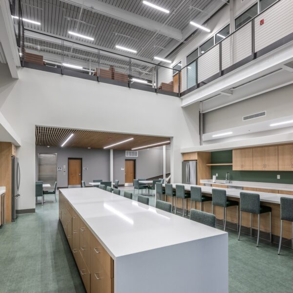 Modern communal kitchen with a large white island, wooden cabinets, green accent walls, barstool seating, and an open loft area with railings above. The space is bright with high ceilings and ample lighting.