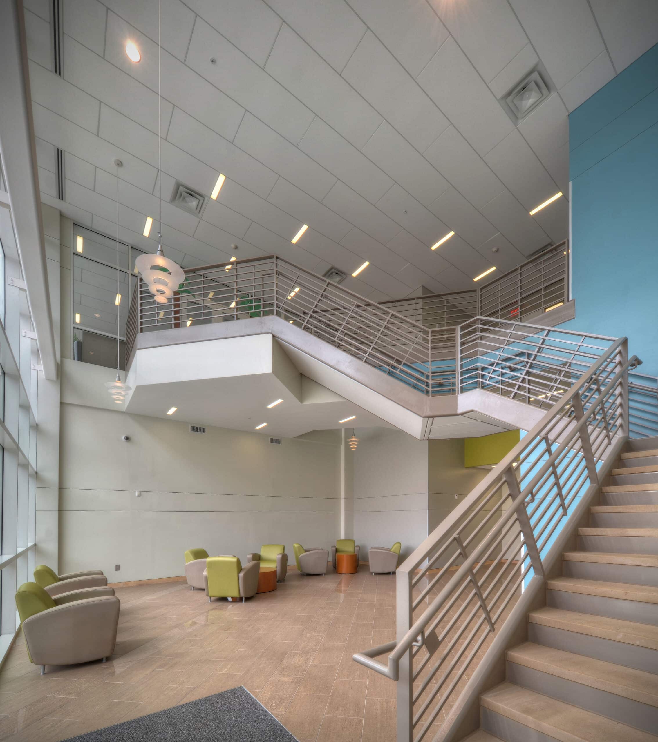 Modern spacious atrium with staircase, metal railing, high ceiling, large windows, and green and beige lounge chairs on beige floor.