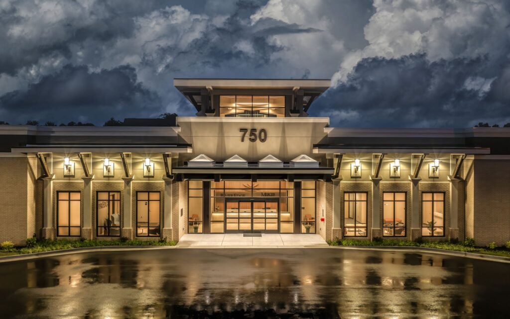 Modern building with large glass entrance labeled 750, lit at night, reflections on wet pavement, dramatic cloudy sky above.
