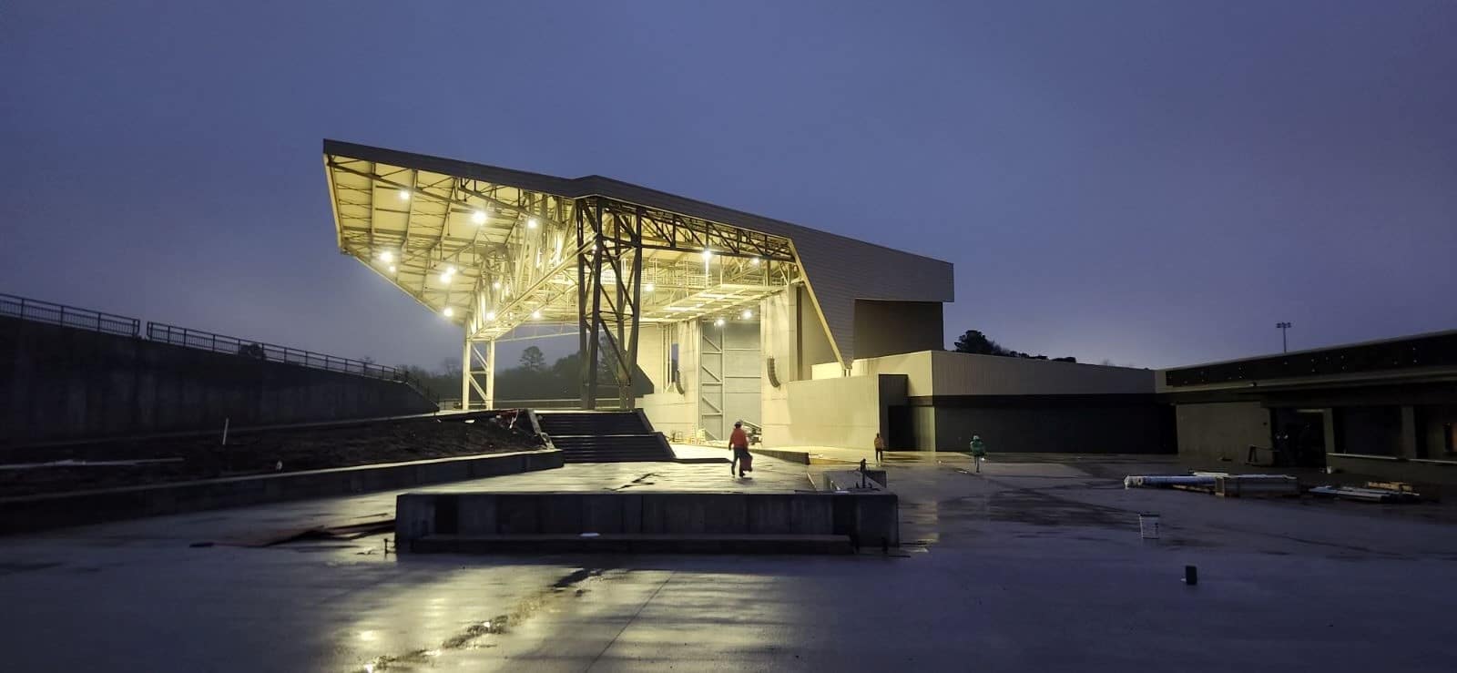 A modern amphitheater with a large angular roof glows at dusk, its lights mirrored on wet ground. A few people stand near the stage.