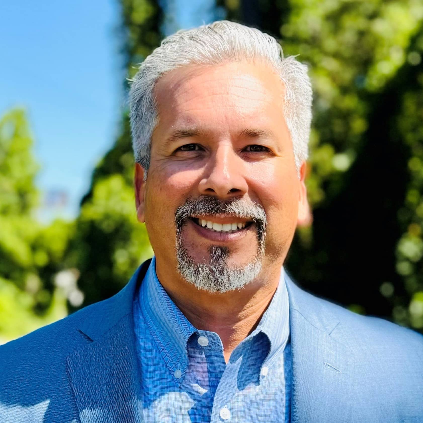 A smiling man with gray hair and a goatee, wearing a blue suit jacket and light blue shirt, stands outside among green trees.