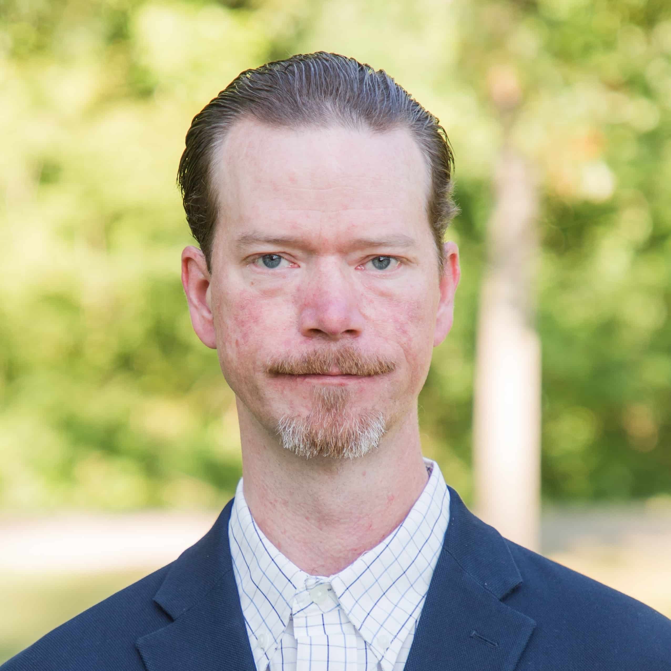 A man with light skin, short brown hair, and a goatee in a blue blazer and white checkered shirt stands outdoors by green trees.