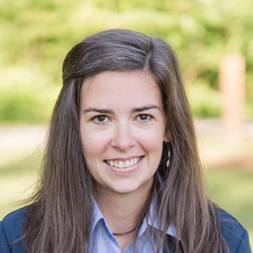 A woman with long brown hair in a blue blazer and light blue shirt smiles outdoors, with greenery and soft sunlight behind her.