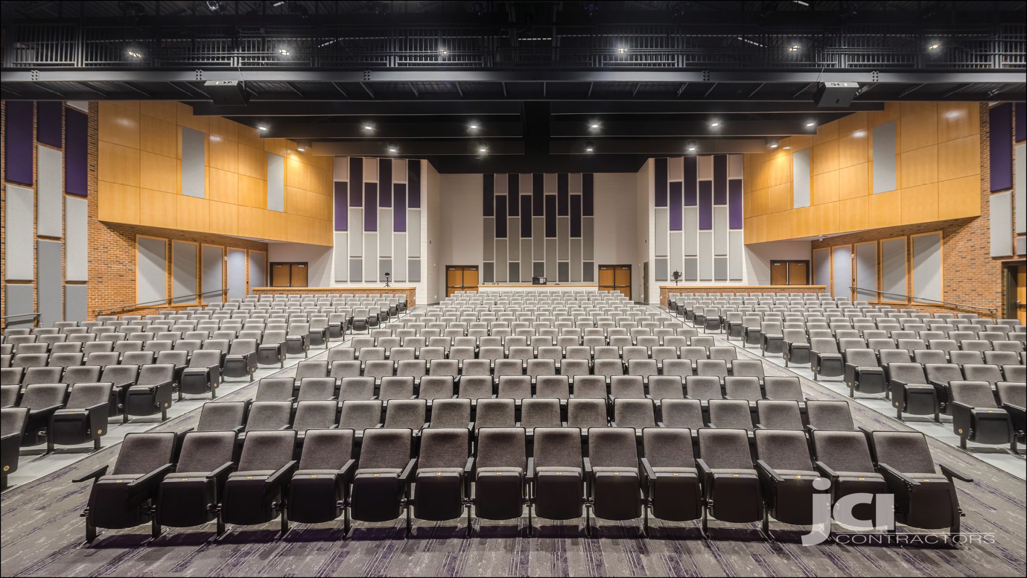 A spacious, empty auditorium with gray seats facing a modern stage, wood and acoustic-paneled walls, and exposed beam ceiling.