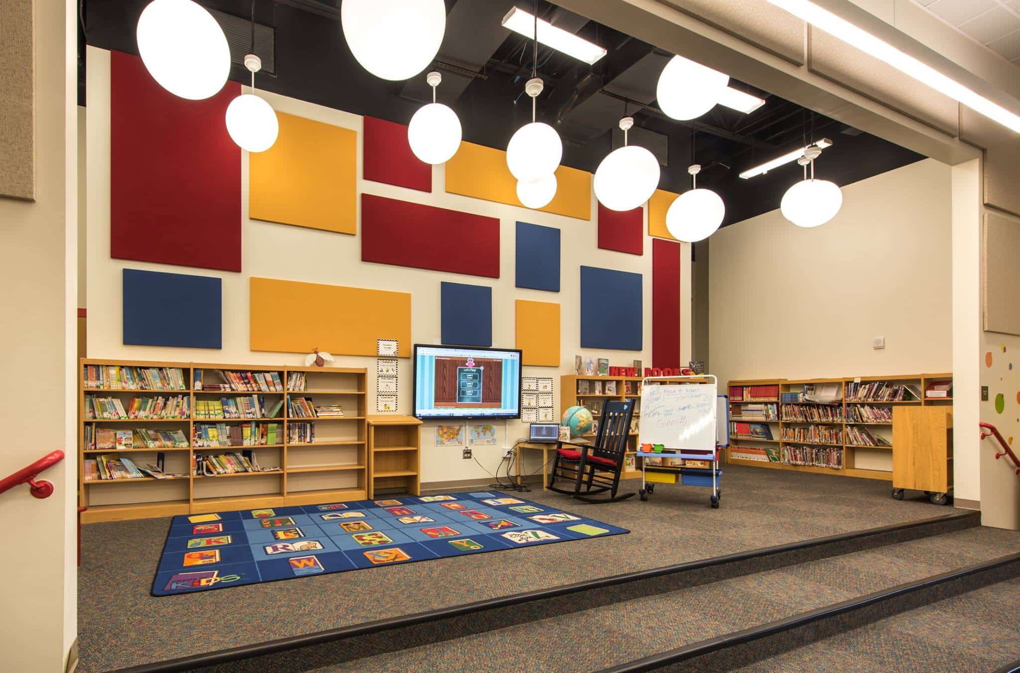 Colorful school library with bookshelves, blue alphabet rug, easel, globe, modern lights, and walls with bold geometric panels.