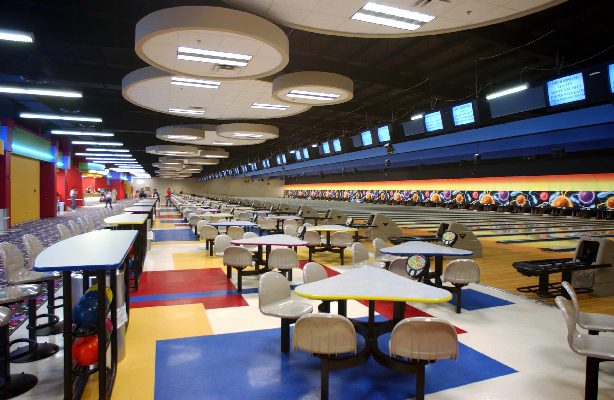 Modern bowling alley with colorful floors, white chairs and tables, several lanes, score screens overhead, and bright lighting.