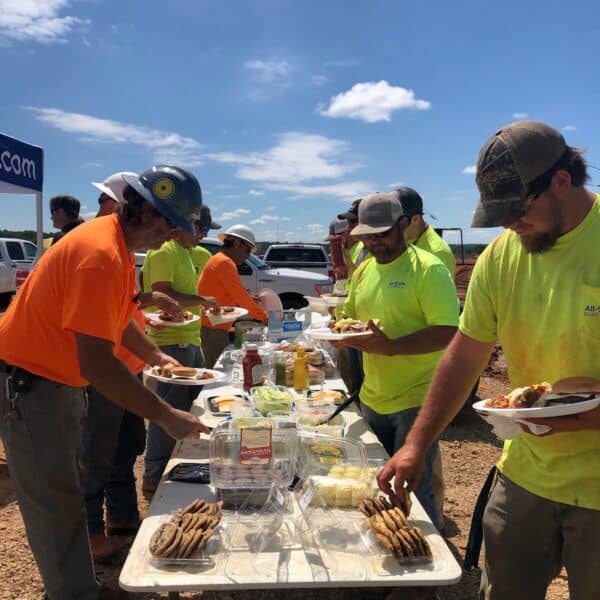 Construction workers in safety vests and bright shirts help themselves to lunch from an outdoor table filled with food on a sunny day.
