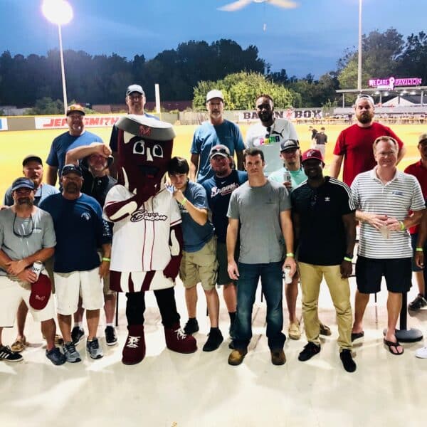 A group poses at a baseball stadium with a maroon and white team mascot in the center; the field and stands are visible behind.