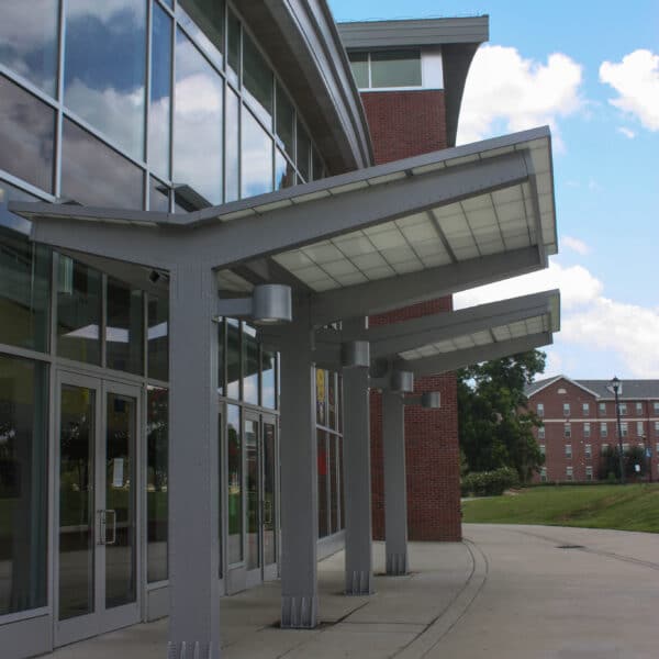A modern building with large glass windows and three metal awnings. Brick building and grassy area behind, partly cloudy sky.