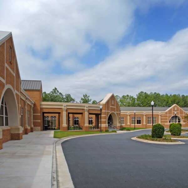 Modern brick school building with arched windows and entrances, mostly empty parking lot, greenery, trees, partly cloudy sky.