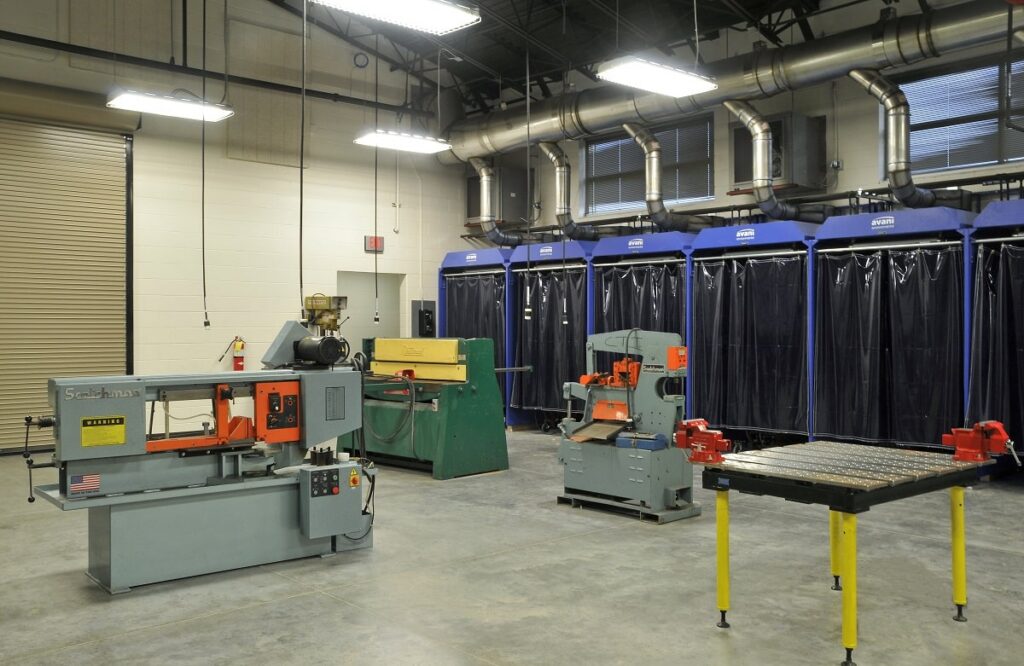 A spacious industrial workshop featuring metalworking machines, welding booths with black curtains, and ventilation pipes running overhead. The floor is uncluttered, and the area is brightly illuminated by ceiling lights.