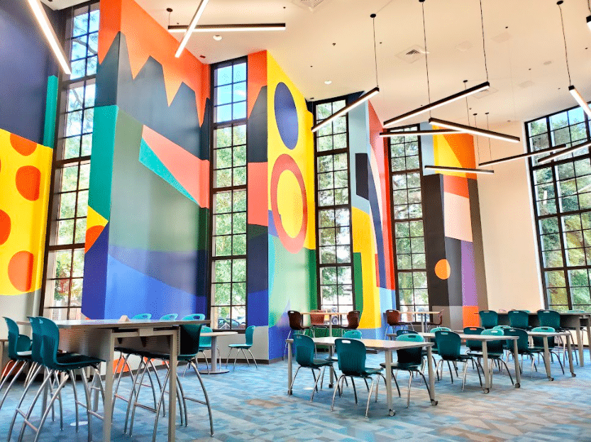 A contemporary study space featuring teal chairs, tables, and expansive windows. The tall walls display vibrant geometric murals, and abstract light fixtures hang from the ceiling.