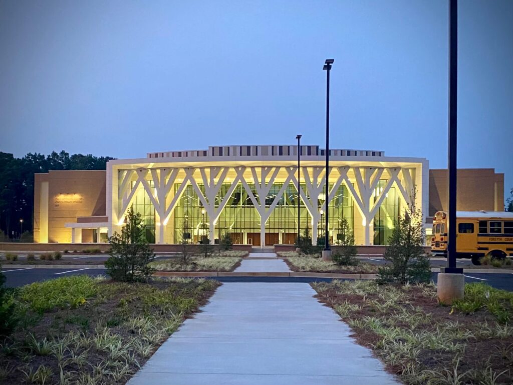 Modern building with large white columns and a glass facade, illuminated at dusk. A sidewalk stretches toward the entrance. Two yellow school buses are parked on the right side. Sparse landscaping and trees surround the area.