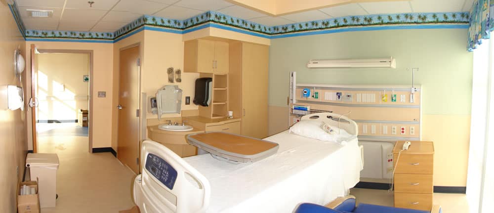 A hospital room featuring a single bed, tray table, sink, cabinets, medical equipment mounted on the wall, and a window dressed with blue curtains. The walls are painted in yellow and green tones with a decorative border running near the ceiling.