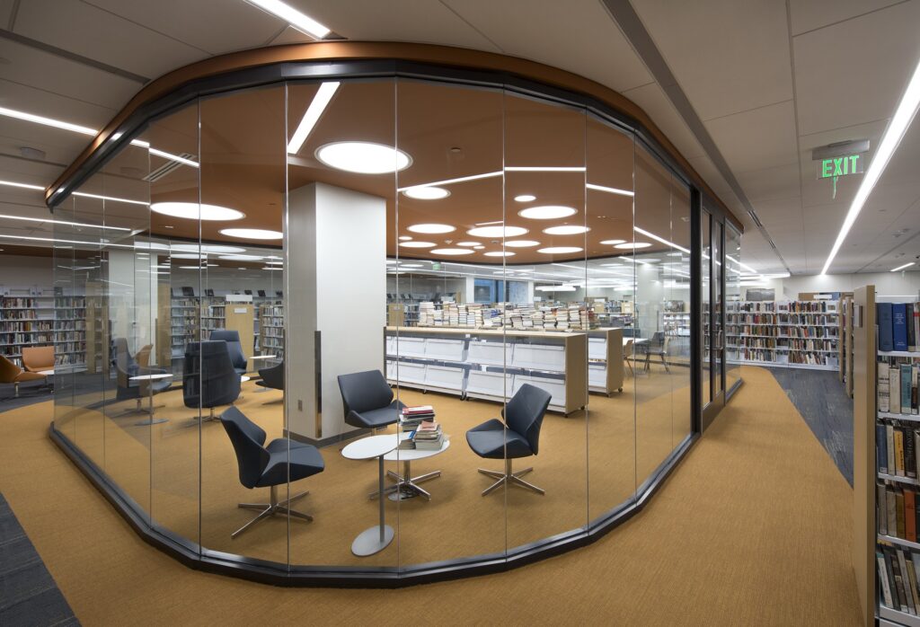Modern library with glass-walled meeting room, four blue chairs around a small table, shelves of books, stacks of books on central tables, and orange-brown carpet throughout. Bright overhead lighting.