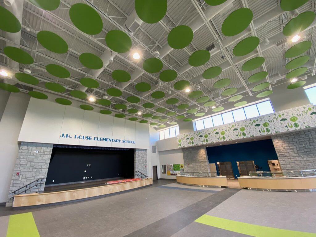 Spacious school cafeteria with green circular ceiling panels, a stage labeled J.H. House Elementary School, large windows, and serving counters. The space is modern and well-lit with natural and artificial light.