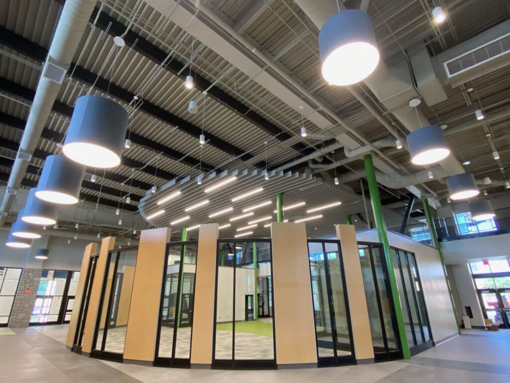 Modern building interior featuring high ceilings, exposed ductwork, circular pendant lights, and a central glass-walled room encased by wooden panels.