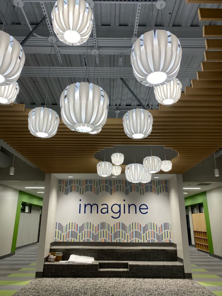 A modern room with white, petal-shaped hanging lights, wood slats on the ceiling, green accents, and the word imagine on the wall above tiered seating with patterned cushions.