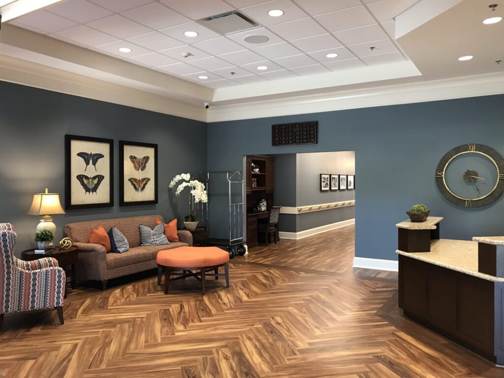 Lobby with wood-patterned floors, blue walls adorned with butterfly art, a cozy sofa, an orange ottoman, an armchair, and a large wall clock. Decorative elements include fresh flowers, lamps, and framed photos. The reception desk is situated on the right side of the space.