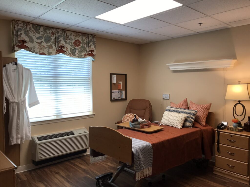 A neatly made bed with orange bedding in a cozy room. A tray rests on the bed. The room includes a window with floral curtains, a hanging robe, lamp, corkboard, and wooden furniture on wood floors.