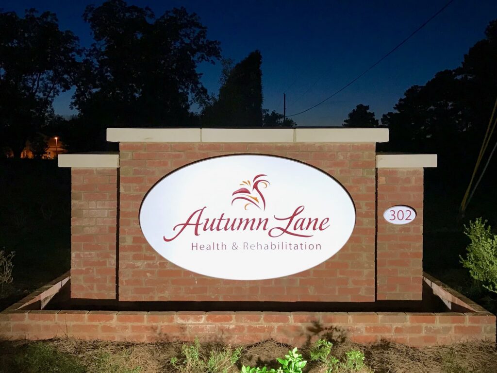 A brick sign for Autumn Lane Health & Rehabilitation illuminated at night, displaying the address number 302 on the right side. Trees and a dark sky appear in the background.