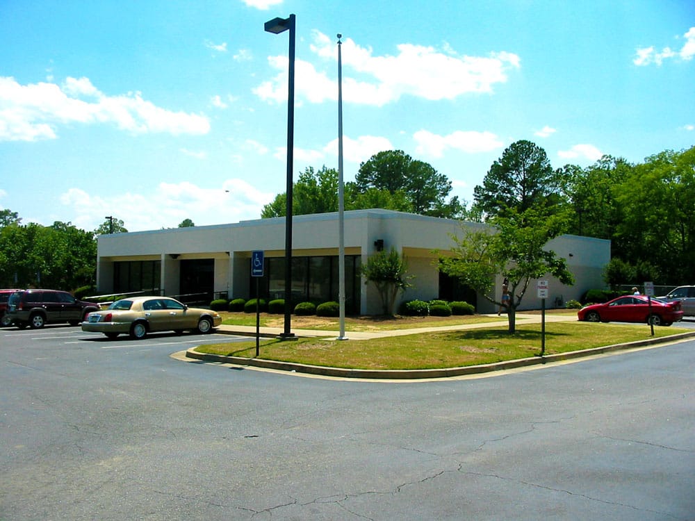 A single-story commercial building featuring large windows stands surrounded by a parking lot where several cars are parked. In the background, trees add greenery beneath a bright, sunny sky.