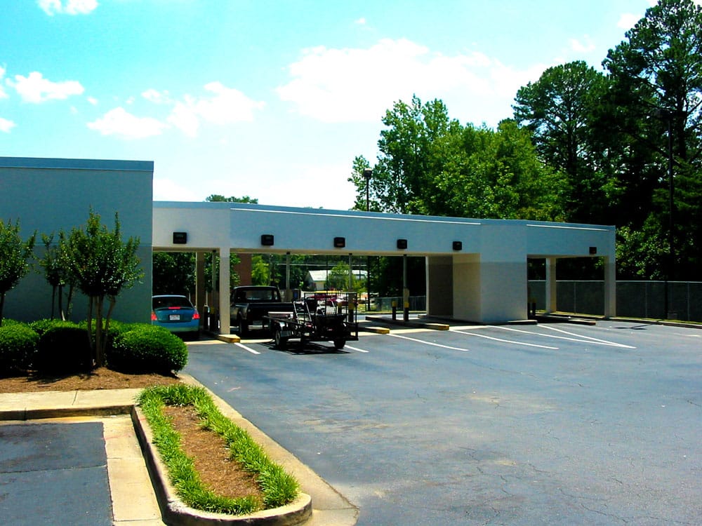 A mostly empty parking lot with a handful of parked cars and some landscaped areas in front of a simple white one-story commercial building, surrounded by trees beneath a bright, partly cloudy sky.