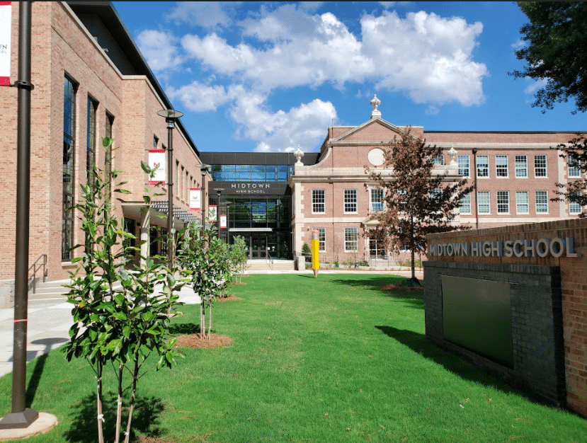 A modern brick school building with a green lawn and small trees in front, a sign reads Midtown High School, and a blue sky with white clouds is above.