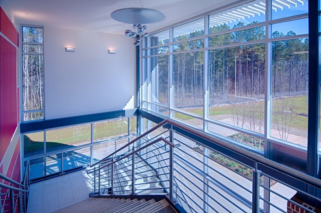A bright, contemporary stairwell with sleek metal railings and expansive windows allowing sunlight to pour in, offering a view of the trees and a winding path beyond.
