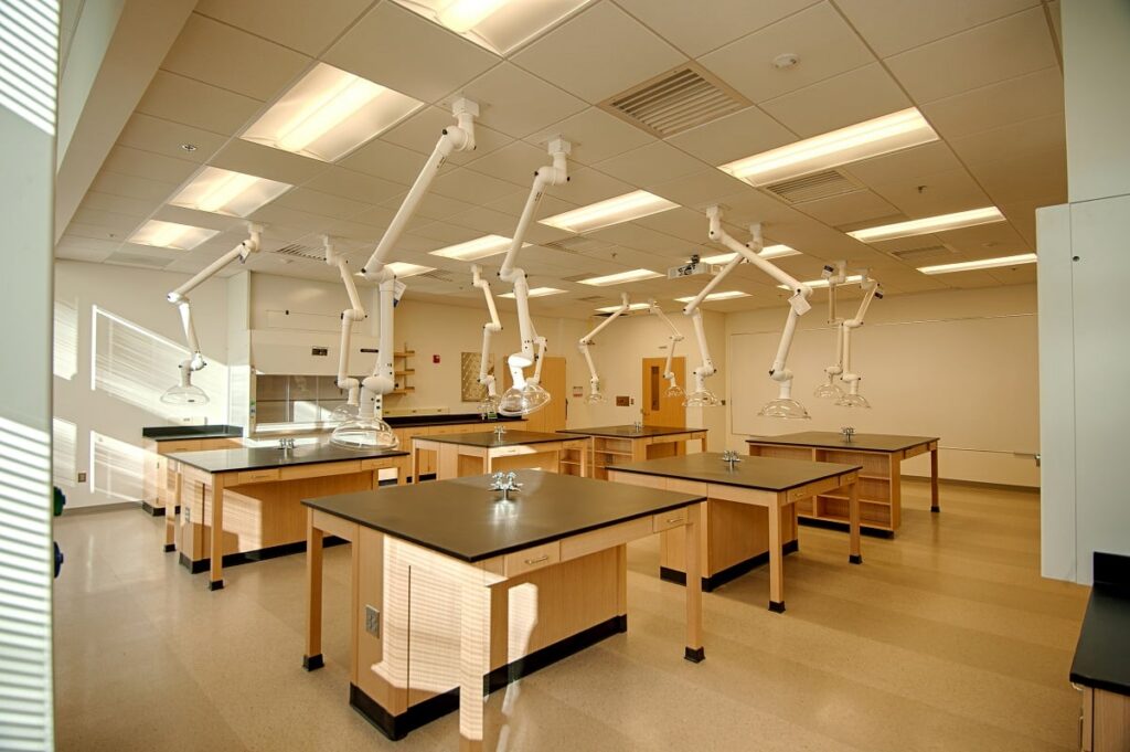 A modern science lab featuring wooden tables with black countertops, ventilation arms, sinks, and bright overhead lighting. The room appears clean and empty, with a fume hood situated in the corner.