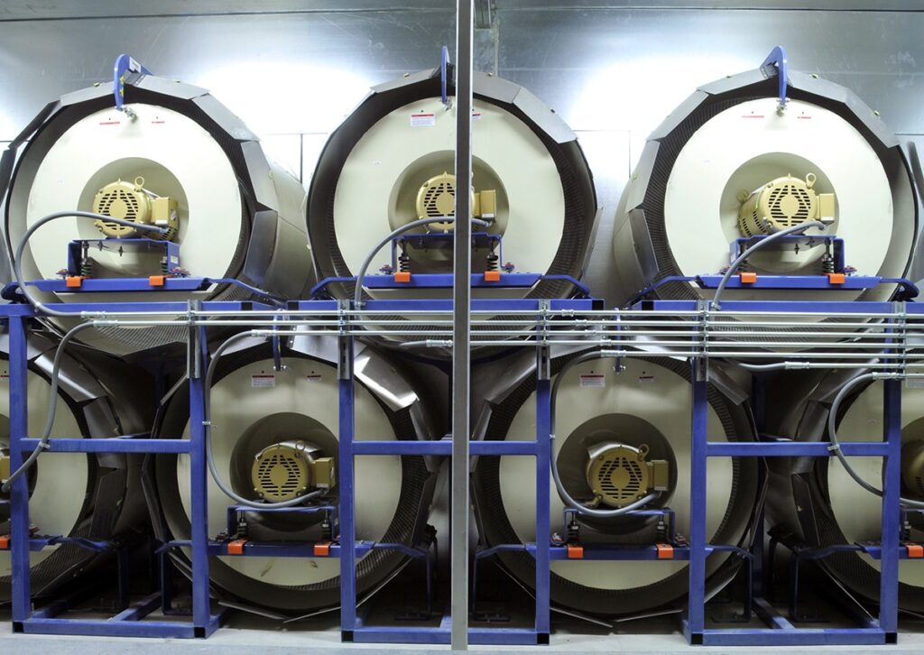 Six large industrial fans are arranged in two rows of three within a blue metal frame, each featuring an electric motor at its center. The setup is located in a pristine, industrial environment.