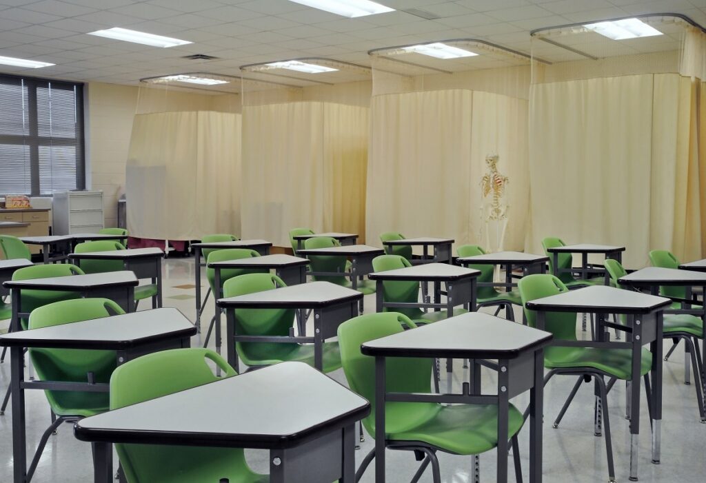 A classroom with green chairs and black desks arranged in rows. Cream curtains divide sections at the back, and a human skeleton model is positioned near the center. Large windows allow daylight to enter.