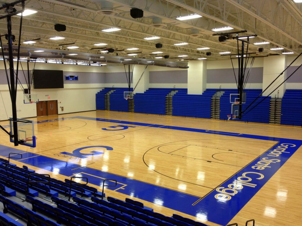 Empty indoor basketball court with polished wooden floors, blue bleachers, multiple basketball hoops, and Gordon State College written on the court. Bright overhead lighting illuminates the gym.
