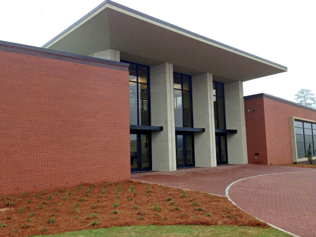 Modern building with large windows, a wide overhanging roof, and red brick walls. Young plants grow in beds near the entrance, and a red brick walkway curves toward the front doors.