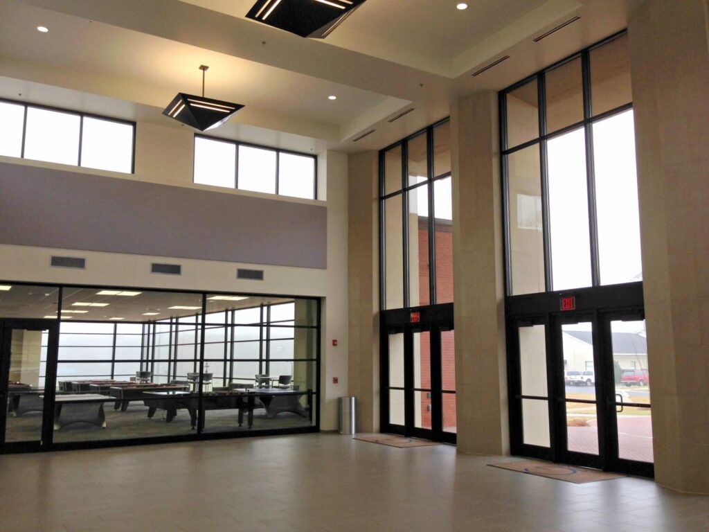 Spacious lobby with tall windows, three glass double doors, and a view into an adjacent room furnished with tables and chairs. Natural light brightens the modern, uncluttered interior.