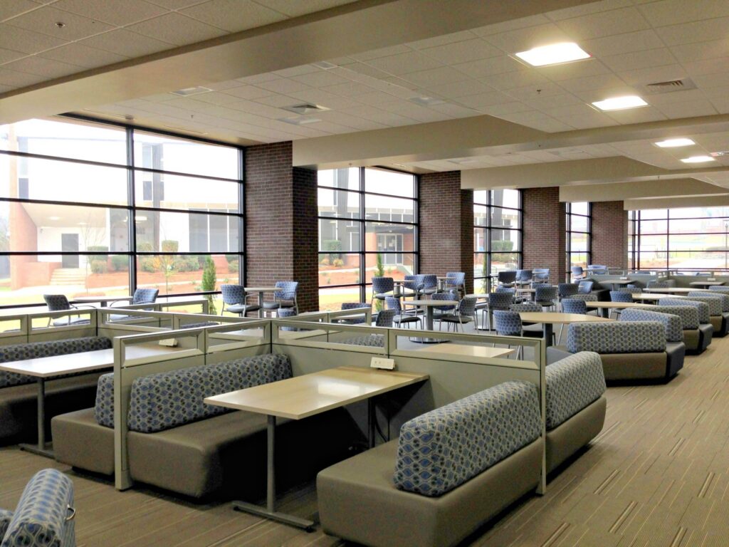 A spacious, modern lounge featuring patterned sofas and square tables, illuminated by large windows that let in plenty of natural light. Additional seating with small tables and chairs is arranged near the windows.