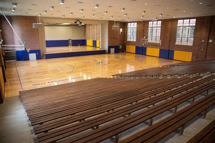 A vacant indoor gymnasium featuring wooden bleachers, a shiny basketball court, blue and yellow wall padding, and large windows that allow ample natural light to enter.