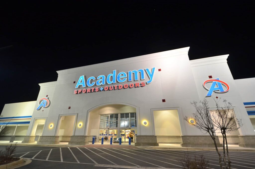 The exterior of an Academy Sports + Outdoors store at night, brightly illuminated with empty parking spaces in front and a clear, dark sky overhead.