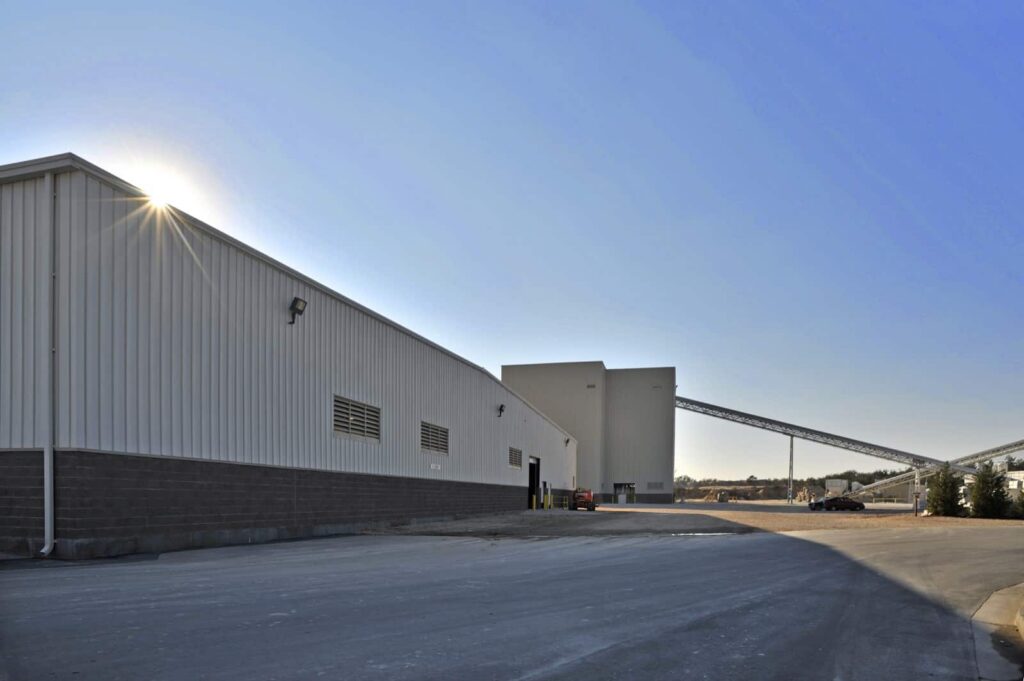 A large industrial building with a metal exterior stands under a clear blue sky, sunlight creating a sunburst effect in one corner. The surroundings are neat, featuring a concrete lot and conveyor belts visible in the background.