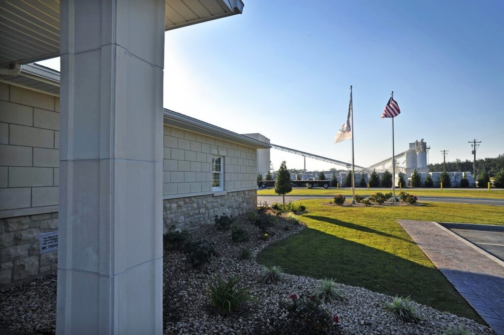 Landscaped grounds feature a building on the left, two flagpoles with the U.S. and another flag, and an industrial facility with silos and conveyors in the background beneath a clear sky.