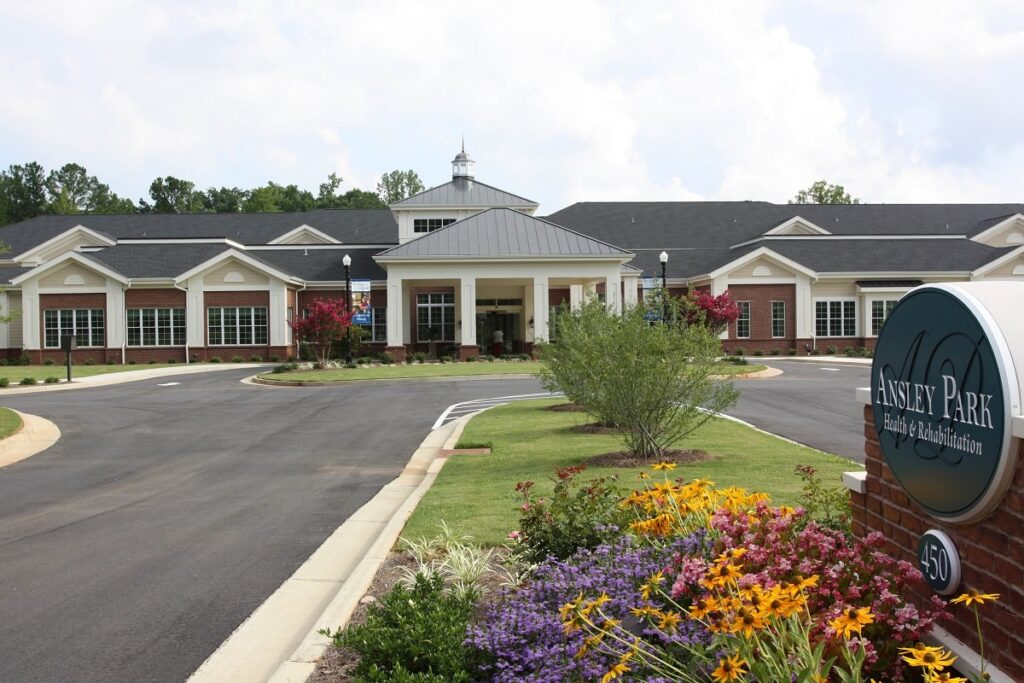 A large, single-story building with a sign reading Ansley Park Health & Rehabilitation is surrounded by colorful flowers, greenery, and a circular driveway under a partly cloudy sky.