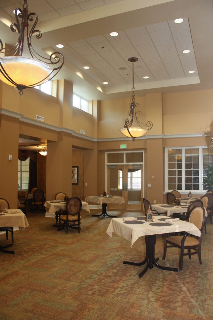 A spacious restaurant with high ceilings, elegant chandeliers, beige walls, and several round tables covered with cream tablecloths and brown cushioned chairs. Large windows let in natural light.