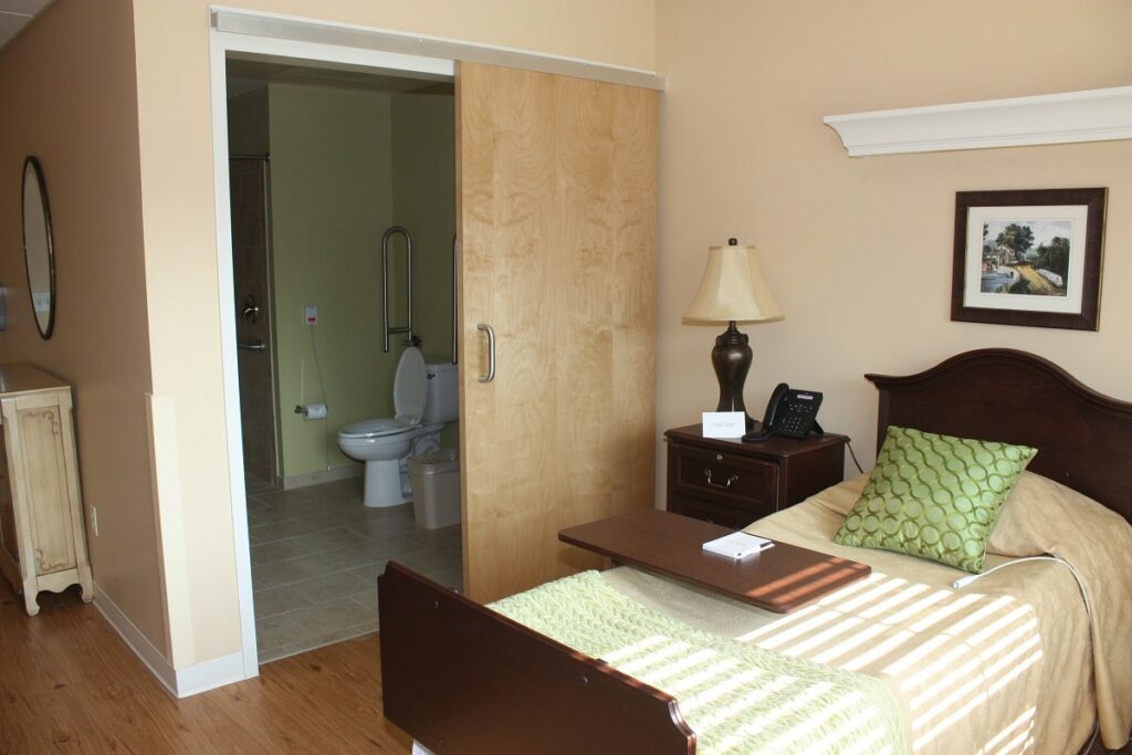 A neatly made single bed with green accents stands beside a nightstand and lamp. An open sliding door reveals a bathroom containing a toilet. Sunlight streams through the window, casting shadows across the bed.
