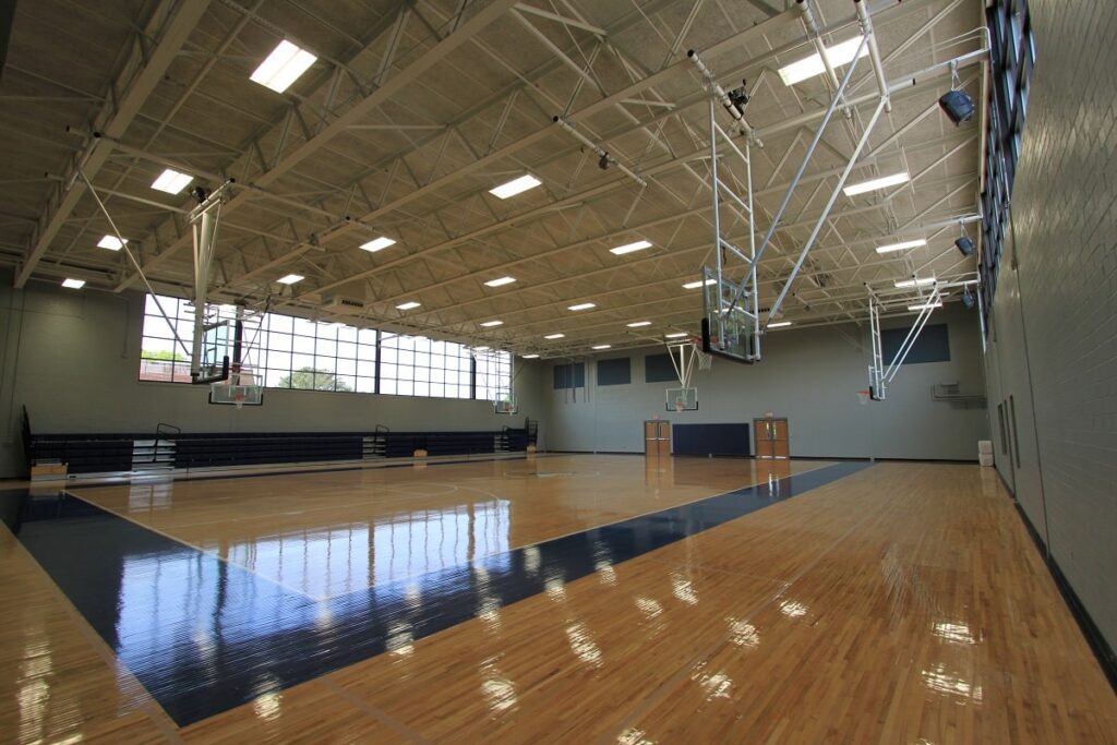 Bright indoor basketball gym with polished wooden floor, high ceilings, large windows, foldable bleachers, and four basketball hoops. The space is empty and well-lit by natural light from the windows.