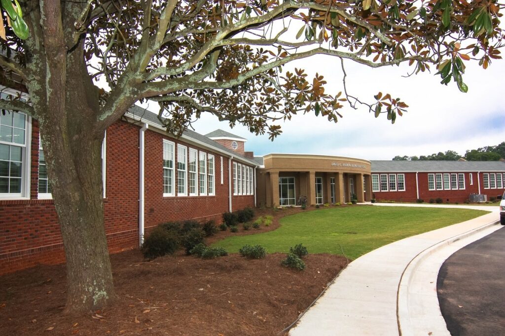A red-brick school building with large windows, a rounded entrance, green lawn, sidewalk, and a tree with broad leaves in the foreground.