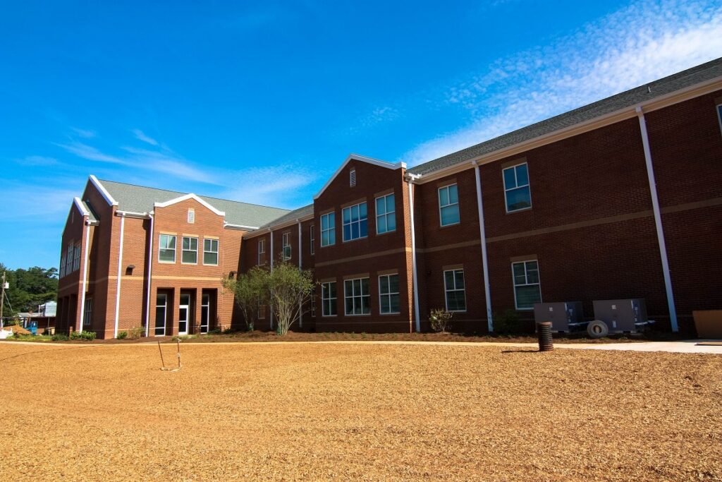 A two-story red brick building with white trim stands beneath a clear blue sky, encircled by a yard covered in dirt and mulch and featuring sparse landscaping.