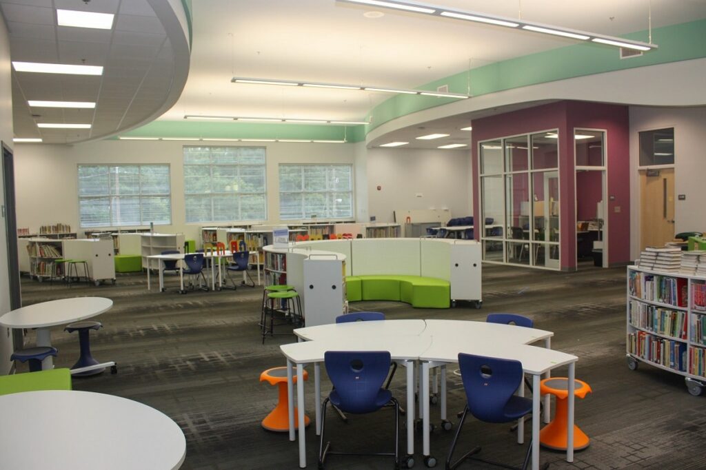 Modern library interior featuring white tables, blue and orange chairs, bookshelves, large windows, and walls painted in green, purple, and white; the area is well-lit and neatly arranged.