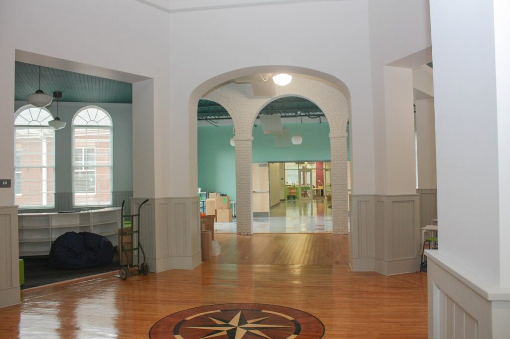 A bright, open hallway featuring wooden floors and white walls, with arched windows along one side. The next room has light blue-green walls, and a compass rose design decorates the floor in the foreground.