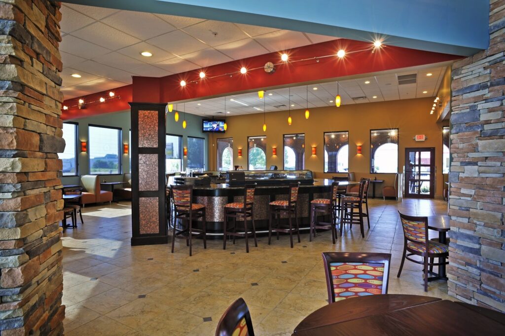 Modern restaurant interior featuring a central bar area, vibrant chairs, stone columns, pendant lights hanging from the ceiling, and expansive windows allowing in natural light. A TV is installed on the far wall above the bar.